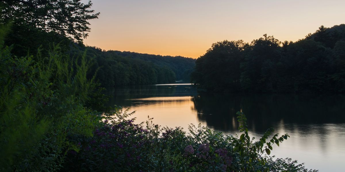 A serene view of a calm lake surrounded by dense forest at Raccoon Creek State Park, Pennsylvania, during sunset, with a clear sky fading from orange to deep blue and reflections of the trees on the water.